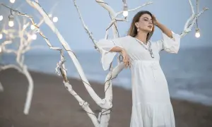 Kobieta w białej sukience opiera się o białe drzewo z lampkami na plaży. Sukienka pięknie układa się, nie podwija się.