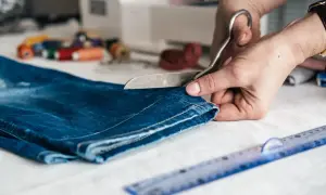 Ręce tną dżinsowe legginsy nożyczkami, przygotowując je do skrócenia. Obok leży linijka i szpulki nici.
