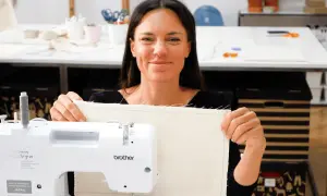 Uśmiechnięta kobieta prezentuje uszyty materiał podczas warsztatów szycia. W tle maszyna do szycia i akcesoria.