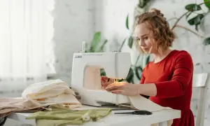 Kobieta w czerwonej sukience szyje na maszynie. Na stole leży dresówka bawełniana i inne tkaniny.