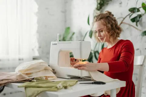 Kobieta w czerwonej sukience szyje na maszynie. Na stole leży dresówka bawełniana i inne tkaniny.