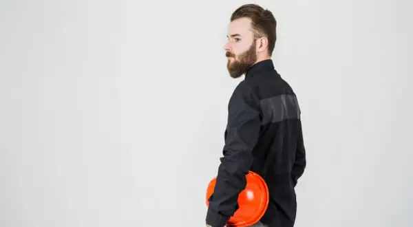 Mężczyzna w czarnej koszuli, z brodą, trzyma pomarańczowy kask. Tkanina na ubrania robocze jest wytrzymała.