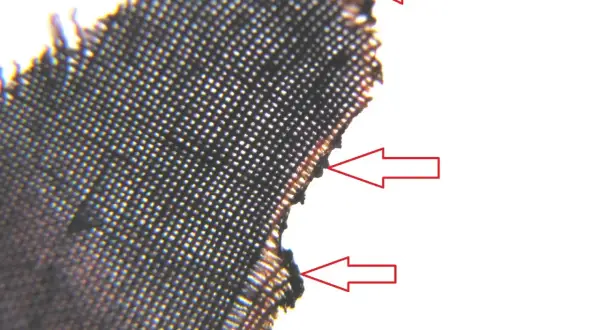 Mikroskopijne zbliżenie na ciemną tkaninę, gdzie czerwone strzałki wskazują na uszkodzone włókna. Czy to pamuk, co to za materiał?