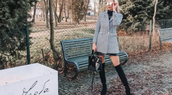 Stylowa kobieta w wełnianym płaszczu w pepitkę, czarnych kozakach i okularach przeciwsłonecznych pozuje w parku.
