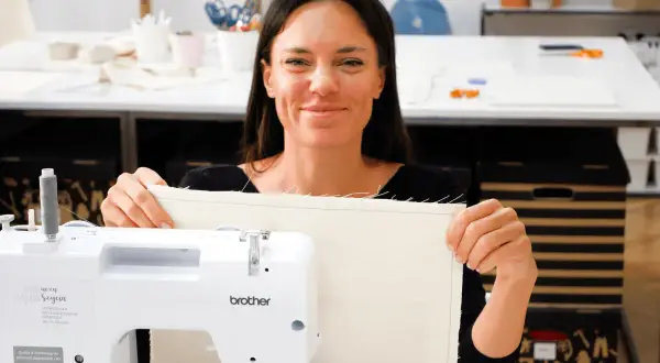 Uśmiechnięta kobieta prezentuje uszyty materiał podczas warsztatów szycia. W tle maszyna do szycia i akcesoria.