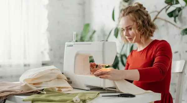Kobieta w czerwonej sukience szyje na maszynie. Na stole leży dresówka bawełniana i inne tkaniny.