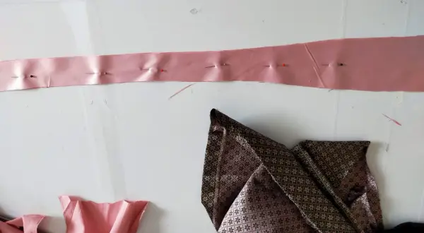 Różowa taśma z agrafkami, przygotowana do poszerzenia sukienki pod pachami. Obok leżą złożone tkaniny.
