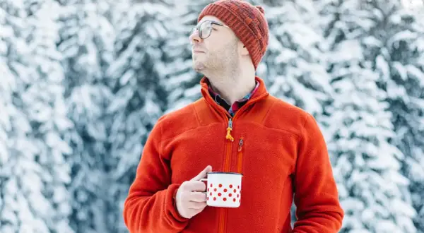 Mężczyzna w pomarańczowym polarze i czapce trzyma kubek w śnieżnym lesie. Polar jest wykonany z miękkiego, ciepłego materiału.