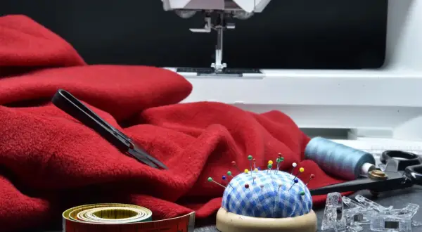 Czerwona, **cienka tkanina** czeka na maszynie do szycia. Obok nożyczki, miarka i poduszeczka z agrafkami.