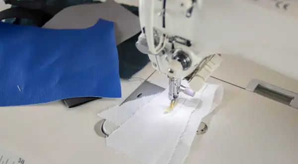 Maszyna do szycia wykonuje stebnowanie na białej tkaninie. Obok leżą skrawki niebieskiej i szarej skóry.
