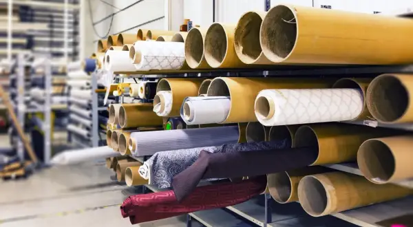 Różne rodzaje materiałów na rolkach, gotowe do użycia w produkcji. Od jasnych wzorzystych tkanin po ciemne, jednolite materiały.