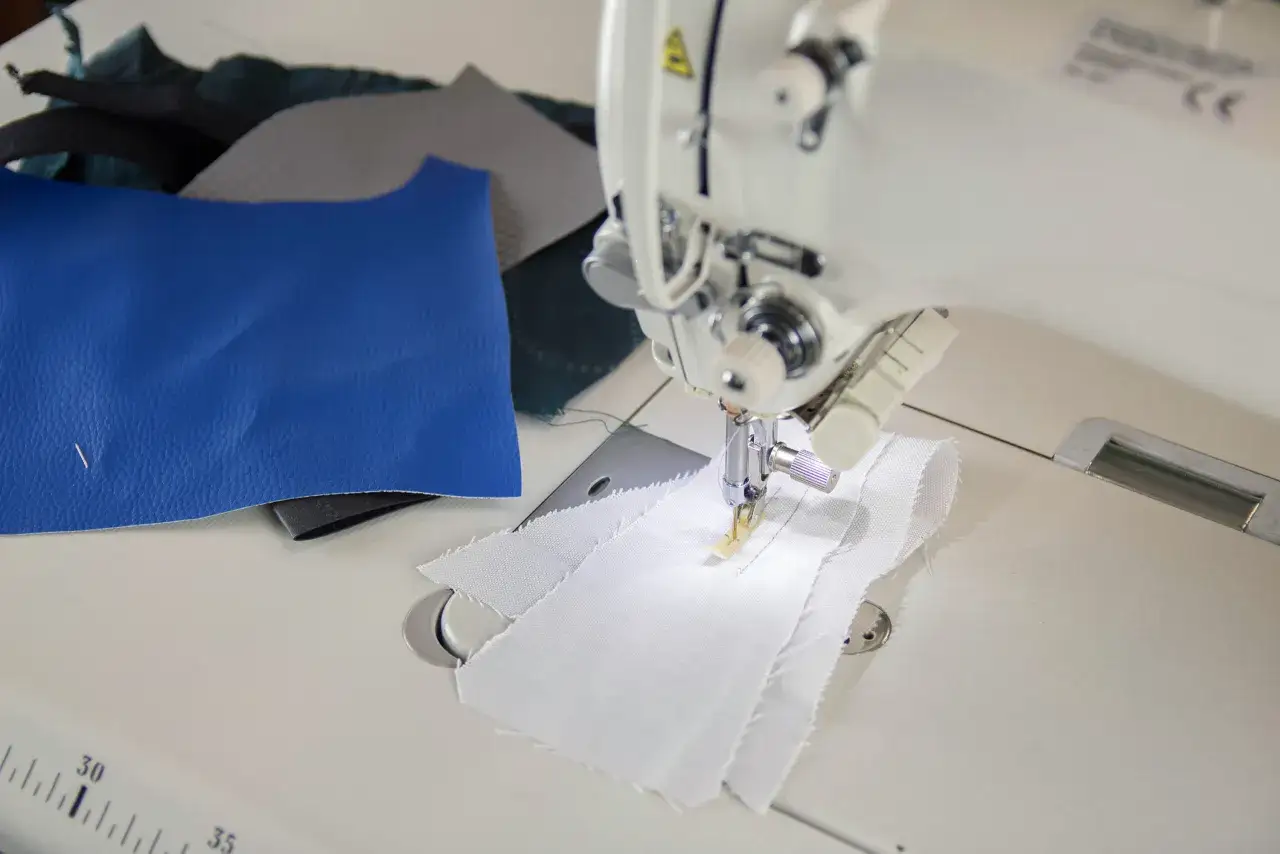 Maszyna do szycia wykonuje stebnowanie na białej tkaninie. Obok leżą pr&oacute;bki niebieskiej sk&oacute;ry i innych materiał&oacute;w.