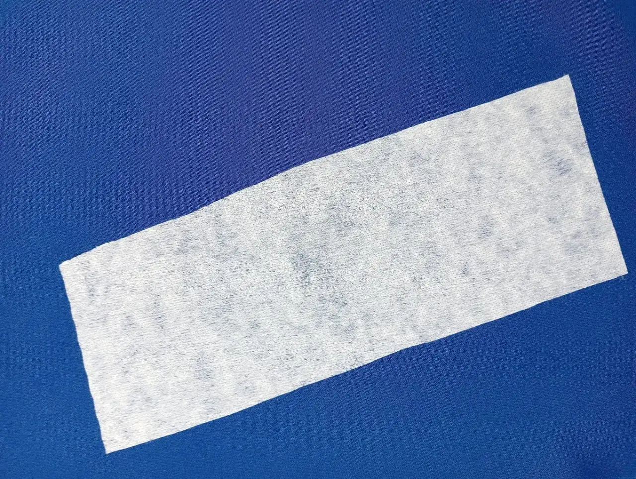 Biały pasek flizeliny (czy fizeliny) na granatowym tle. Materiał jest lekko prześwitujący i ma widoczną strukturę włókien.