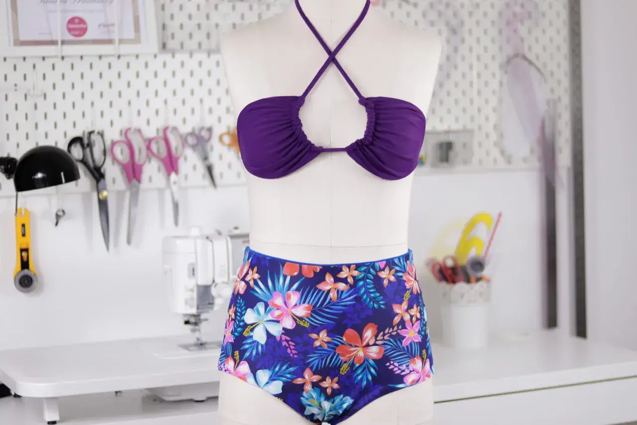 Jak uszyć str&oacute;j kąpielowy? Fioletowy top bikini i figi w tropikalne wzory na manekinie, obok maszyna do szycia i akcesoria krawieckie.