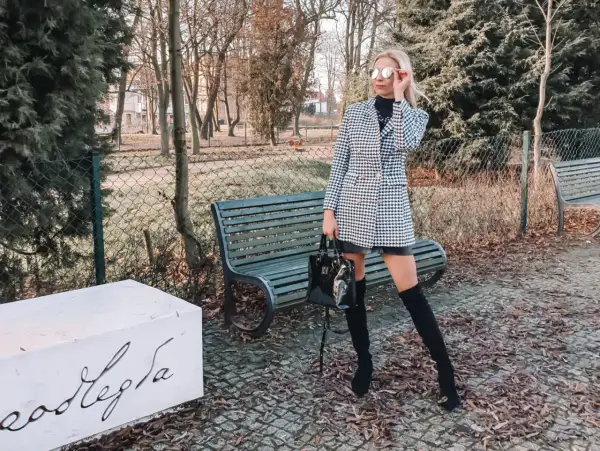 Stylowa kobieta w wełnianym płaszczu w pepitkę, czarnych kozakach i okularach przeciwsłonecznych pozuje w parku.